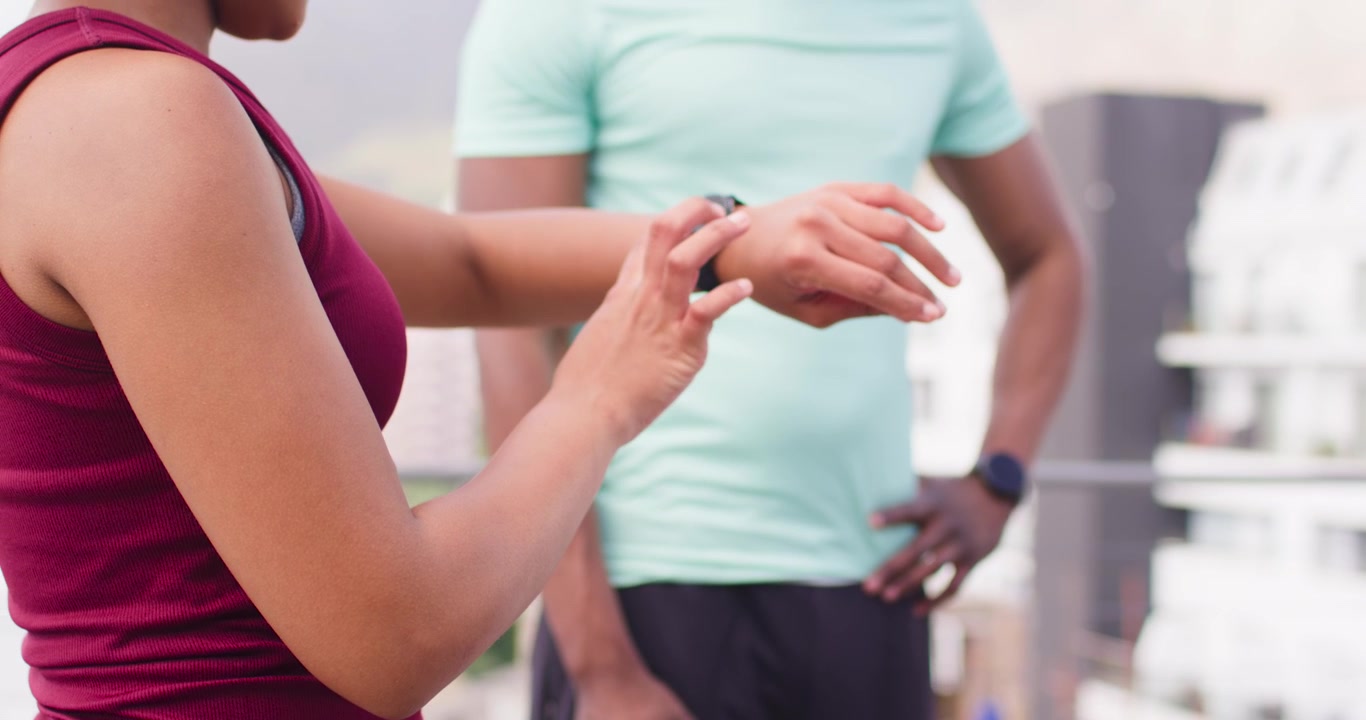 Afro-Amerikaanse fitnesspartners op dakterras, vrouw in tanktop stelt smartwatch in en start training.