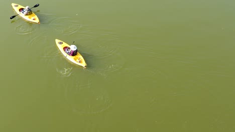 a close-up aerial view of two people slowly kayaking from the bottom middle to the top left of the screen on a calm, sunny day