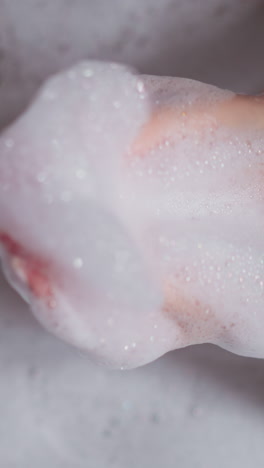 mujer juega con espuma esponjosa en la bañera de primer plano. dama sostiene un puñado de burbujas de jabón descansando en el baño acogedor. mujer se relaja durante la rutina higiénica