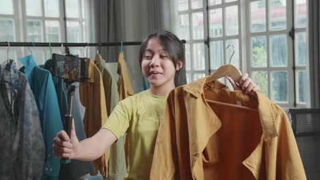 asian young girl online seller recommending a shirt and recording video by mobile phone while selling clothes at home
