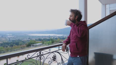 man standing up from the chair and looking towards the horizon on a balcony with a stunning view while holding a cup of coffee