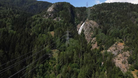 los cables eléctricos pasan por el bosque verde en las montañas nevadas con cascada en el fondo en italia