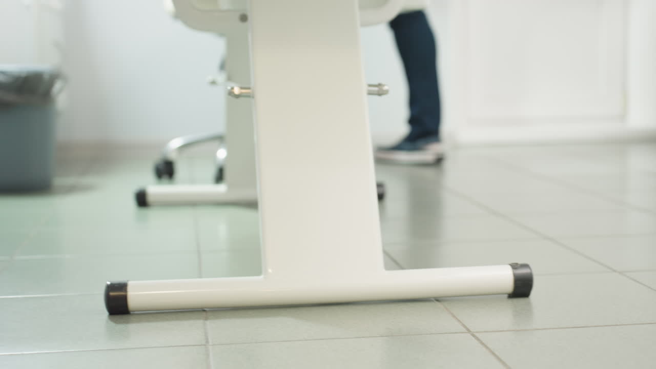 Patient wearing plastic shoe cover walks into bright white clinic room with tiled floor, approaching massage chair alongside therapist to begin treatment, highlighting hygiene and safety measures