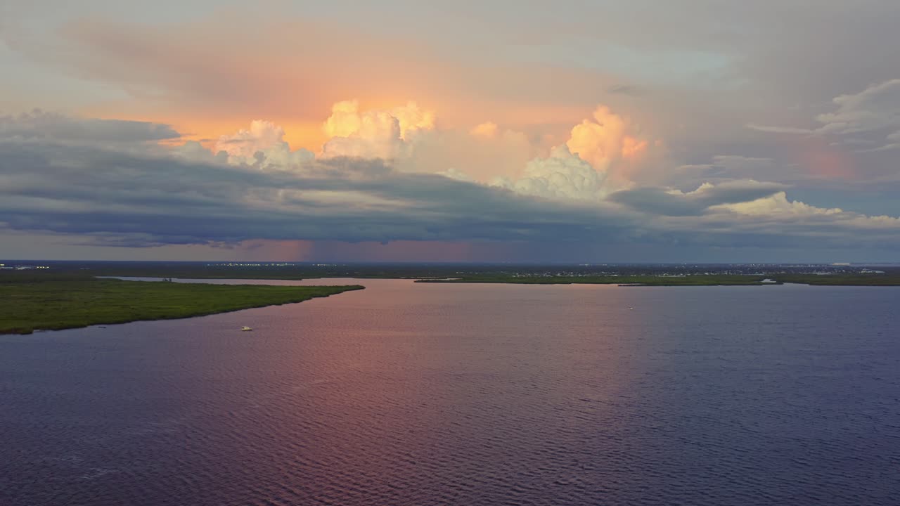 Golden and pink light from the setting sun spills across a calm bay, mirroring dramatic cumulus clouds that tower above the horizon in a tranquil evening scene