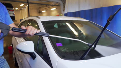 Trabajador-Rociando-Agua-Y-Lavando-El-Parabrisas,-De-Un-Automóvil-Todoterreno-Blanco,-En-Un-Lavadero-De-Automóviles,-En-Suecia