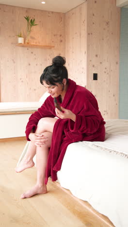 vertical video: woman in red robe applying lotion on legs, relaxing at home
