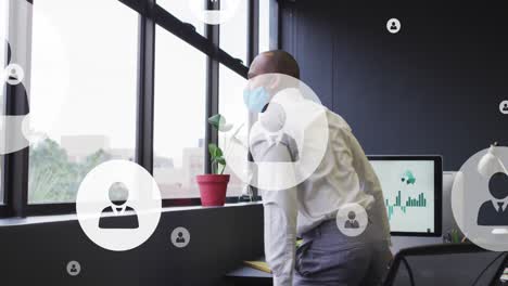 animación de iconos de perfil flotando sobre un hombre biracial con máscara facial mirando por la ventana en la oficina