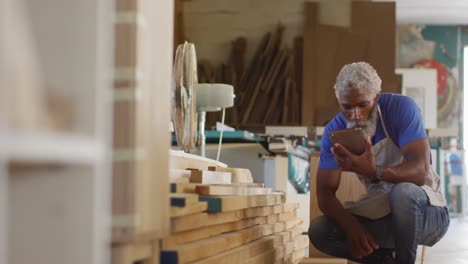 carpintero afroamericano con tableta digital eligiendo un tablón de madera en una carpintería
