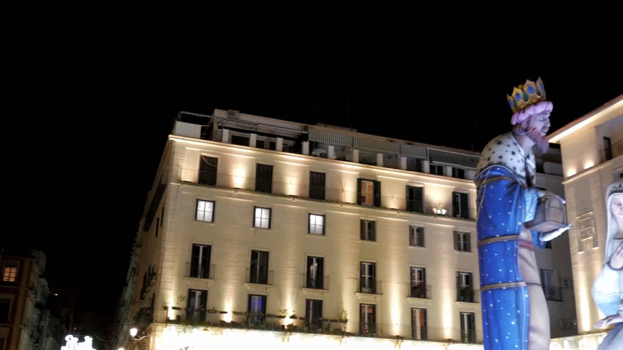 Pan left of Giant Nativity figures in the city at night around the buildings and the flags in the town hall of Alicante, Spain.
