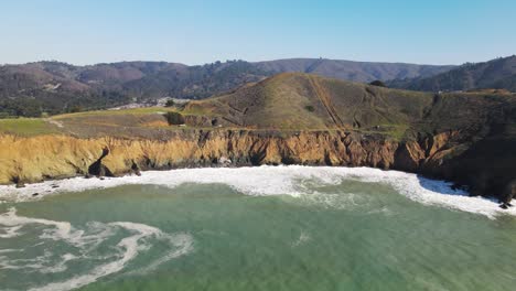 aerial view of mountains parallax at mori point beach coastal cliffs with turquoise waves hitting the shore, drone pan left shot