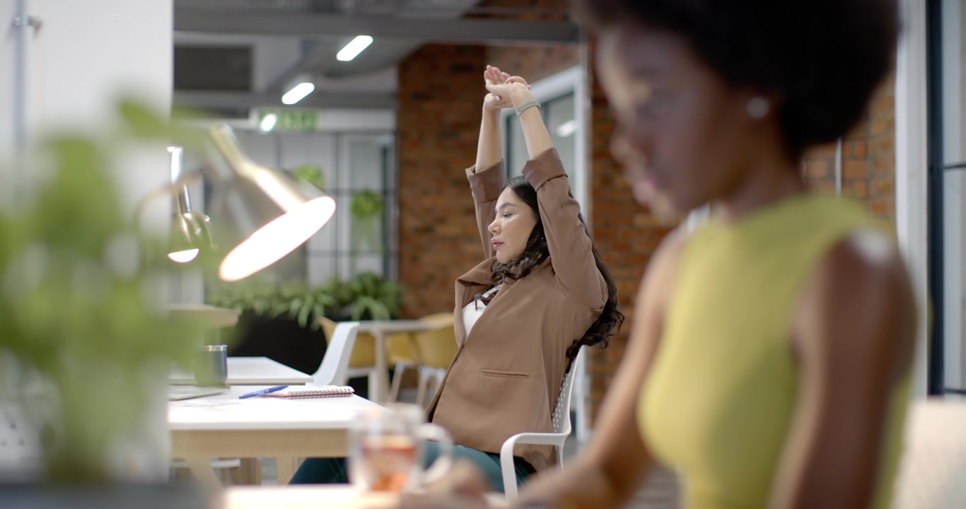 African woman typing on laptop, lens pulling back revealing Hispanic woman stretching, copy space