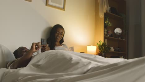 young couple at home at night lying in bed looking at mobile phones together 1