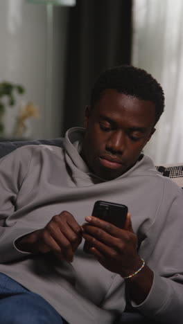 vertical video of unhappy and depressed young man sitting on sofa at home messaging or browsing on mobile phone shaking head