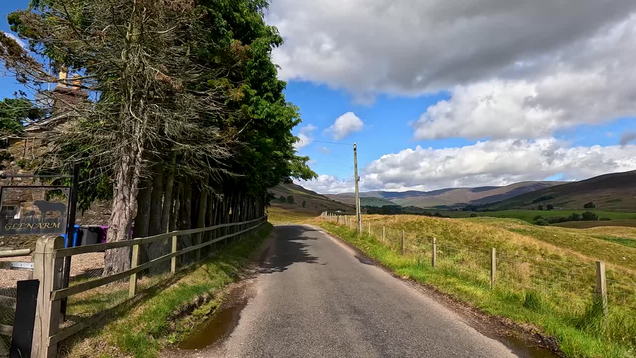 Vehicle drives along narrow rural road past fields, trees, and stone buildings under bright sky