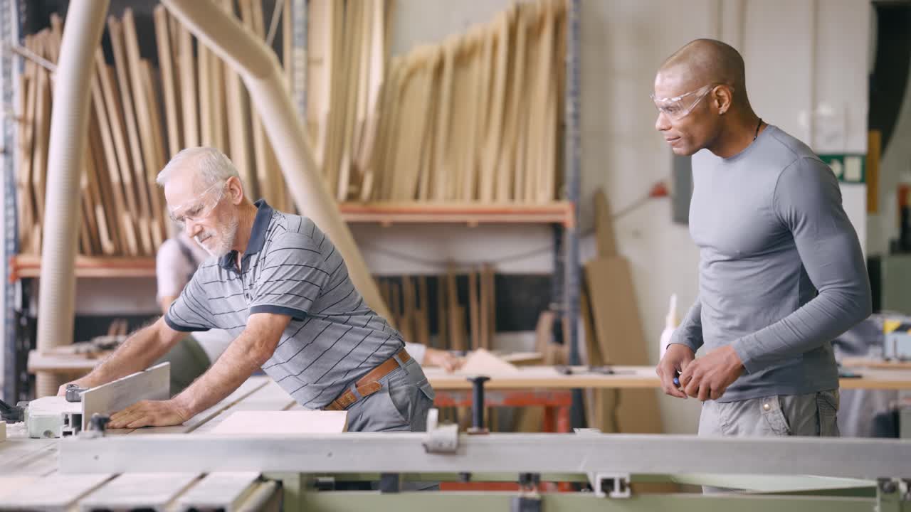 Older Carpenter Working with Wood on Machine While Younger Man Observes