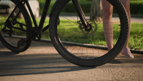 close-up of someone's legs as they gently kick a bicycle tire, with sunlight illuminating the scene and blurred greenery in the background