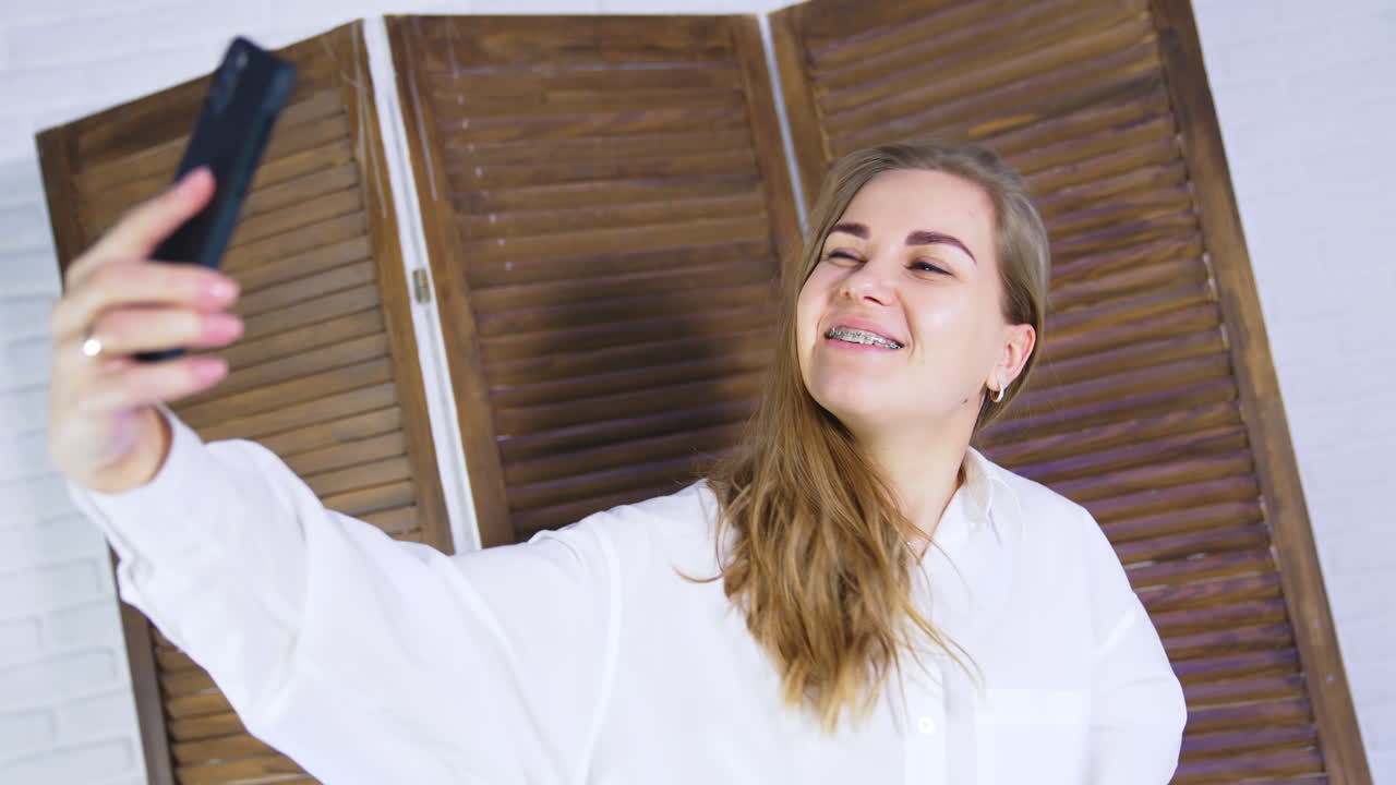 Happy smiling lady posing in front of phone. Cheerful woman making faces and taking selfie on her smartphone. Wooden door backdrop.