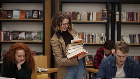 Imágenes-En-Cámara-Lenta-De-Una-Chica-Con-Chaqueta-Marrón-Y-Gafas-Caminando-Por-Una-Fila-Con-Un-Montón-De-Libros-Que-Sacó-Del-Estante-Y-Comenzando-A-Leer-El-Libro.-Otros-Estudiantes-Sentados,-Estudiando-En-La-Universidad,-Biblioteca-Universitaria.-Vista-Frontal