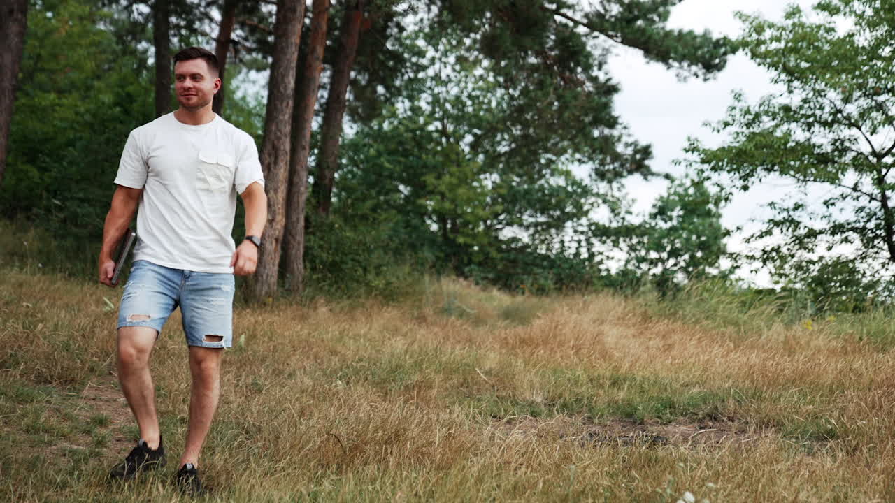 Tall muscular Caucasian man wearing white t-shirt and jeans shorts walks in the nature. Man holds a laptop, sits down into folding chair to work remotely.