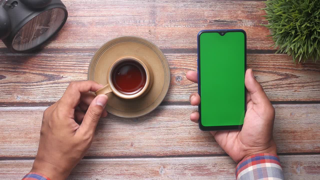 persona sosteniendo un teléfono inteligente y una taza de té en una mesa de madera