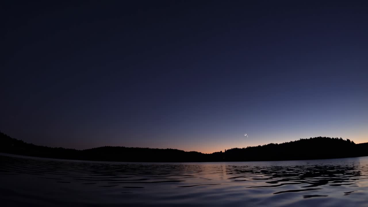 Um vídeo sereno do crepúsculo captura uma vista de ângulo baixo de um lago calmo com árvores em silhueta.