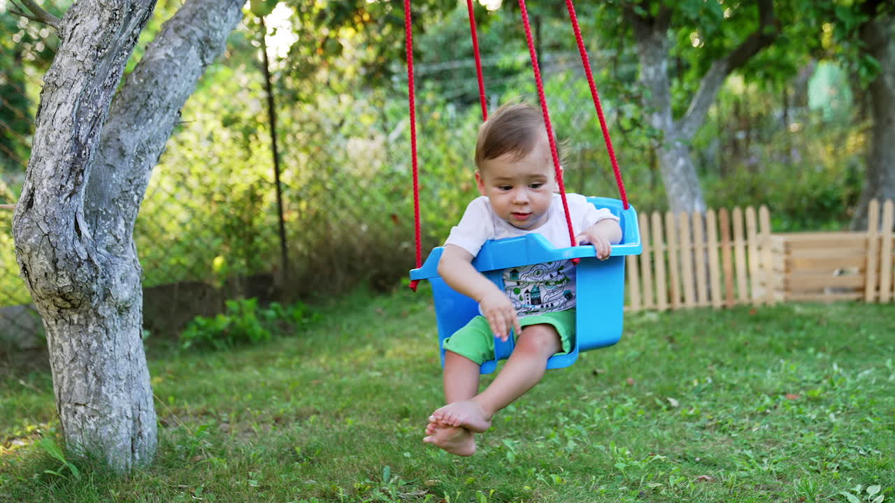 Lovely barefoot Caucasian boy swinging in a blue swing hanging from a tree. Toddler waving hand happily swaying. Garden background.