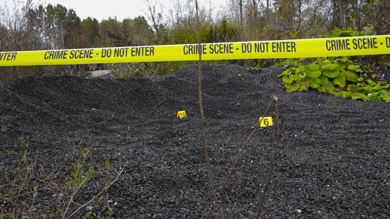 Yellow police line crime scene forensic investigation tape hanging in front of a outdoor nature area with numbered police crime scene markers next to evidence on dark colored gravel. Forest background