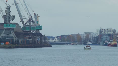 Small-white-coastal-fishing-boat-leaves-the-Port-Of-Liepaja,-overcast-day,-fisherman-ready-to-catch-a-fish-in-a-Baltic-sea,-local-business,-port-cranes-in-background,-wide-distant-shot