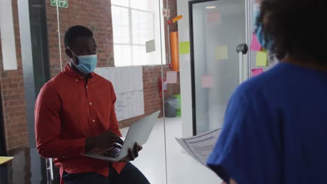 African-american-businessman-with-laptop-discussing-memo-notes-with-colleague-wearing-face-mask