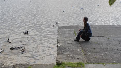 Blonde woman kneels to feed ducks and swans by the river, space for text