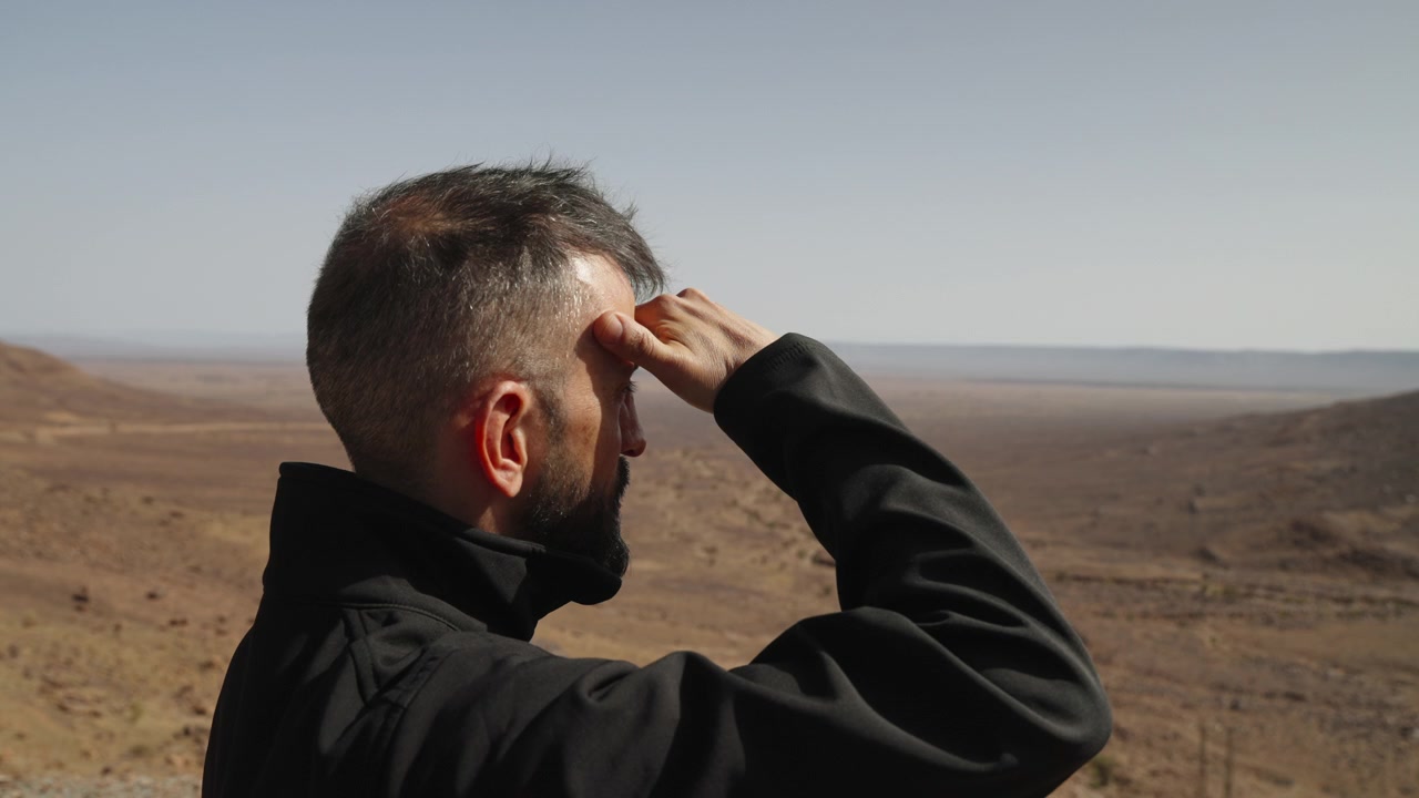 Uomo barbuto in giacca nera che si ripara gli occhi con la mano, osservando una vasta pianura desertica che si estende fino all'orizzonte nel Marocco meridionale.