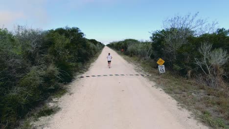 Atleta-Femenina-Corriendo,-Corredora-En-Zona-Rural,-Drone-Disparado-Desde-Atrás,-Mujer-Entrenando-Para-Competir-En-Una-Carretera-Desierta-Entre-Arbustos-Verdes