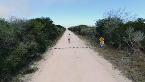 atleta femenina corriendo, corredora en una zona rural, drone disparado por detrás, mujer entrenando para la competencia en una carretera desierta entre arbustos verdes