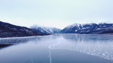 Frozen-lake-with-snow-capped-mountains-in-the-background-and-an-overcast-sky,-dolly