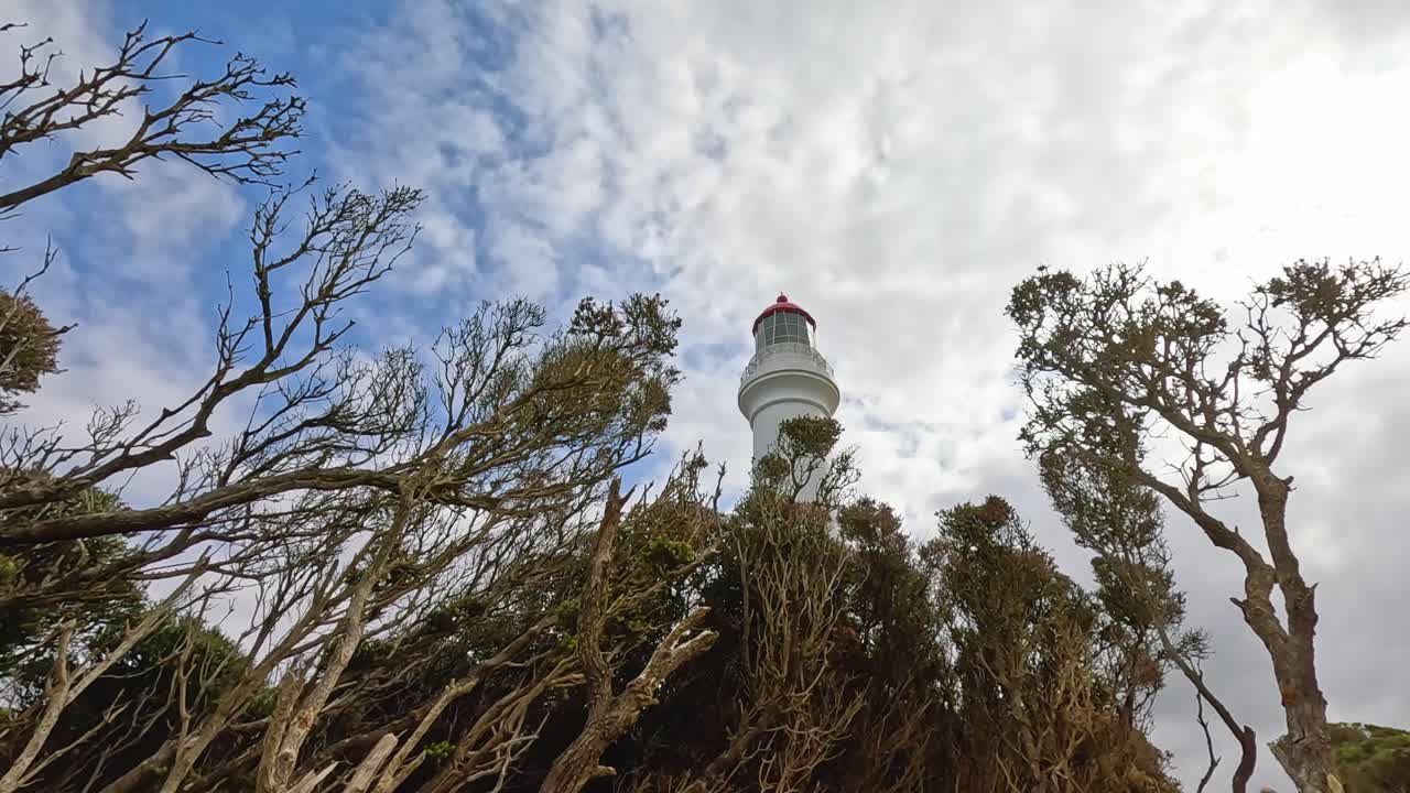 A lighthouse stands tall among trees, framed by a partly cloudy sky. The scene captures natural beauty and architectural elegance