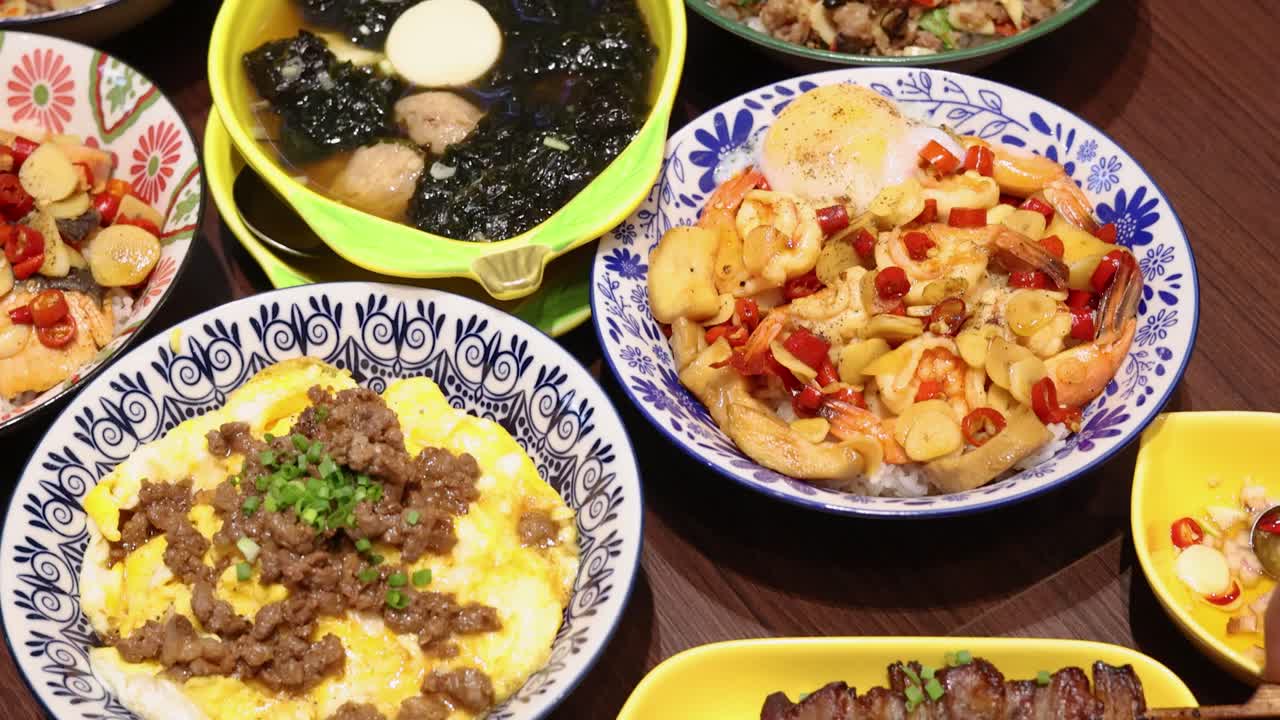 Overhead view of a vibrant Thai rice meal with multiple colorful dishes, including soups, stir-fries, and grilled skewers, under bright indoor lighting with smooth camera movement
