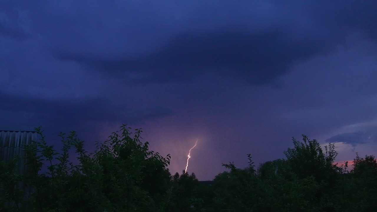 Dark blue skies in the rural countryside. Tops of trees and roof of the cottage house. Sudden lightning breaking the skies.