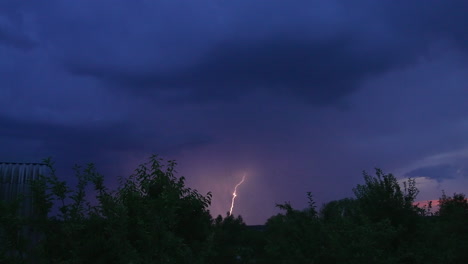 dark blue skies in the rural countryside. tops of trees and roof of the cottage house. sudden lightning breaking the skies.