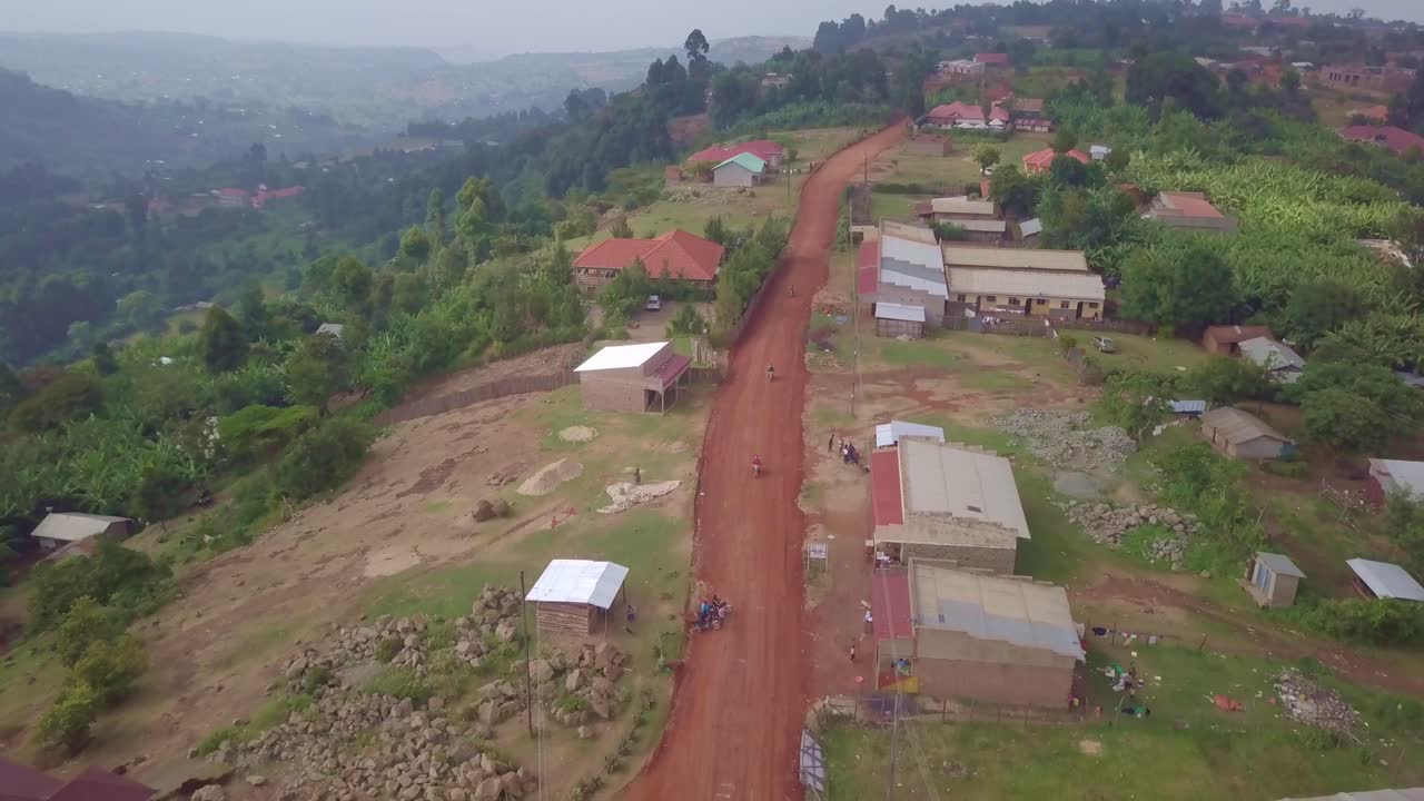 Aerial pull-away shot captures motorcycles traveling a red unpaved road through the hilly townscape of Kapchorwa, Uganda, with scattered homes and vegetation surrounding the elevated terrain