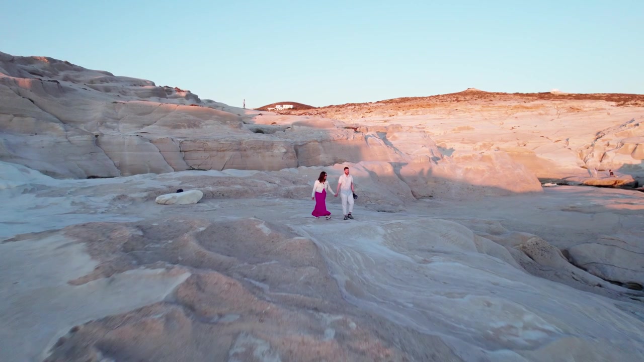 Ripresa aerea con drone in allontanamento di una coppia mano nella mano che cammina sulle rocce vulcaniche bianche dall'aspetto lunare di Sarakiniko, a Milos, in Grecia, durante l'ora dorata