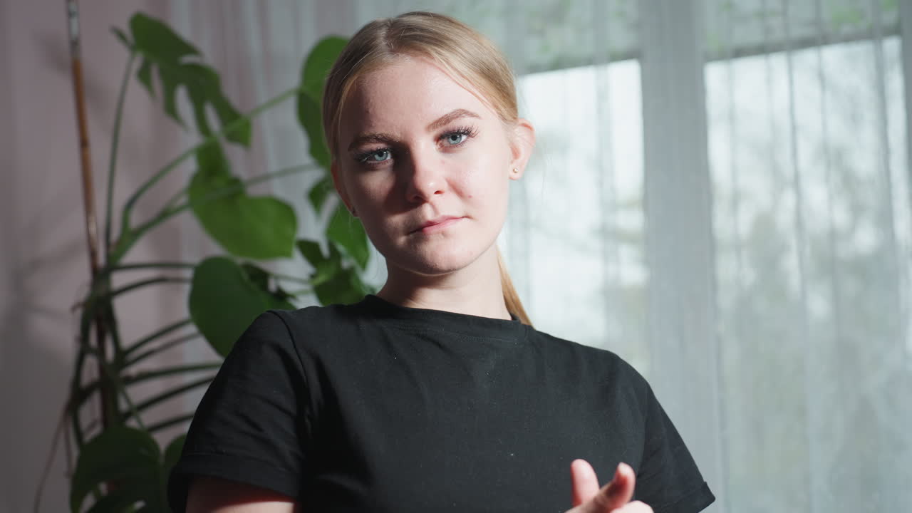 Close up of massage therapist in black shirt looking directly at camera while gently rubbing hands, standing in front of leafy indoor plant and soft curtain backdrop with natural daylight ambiance