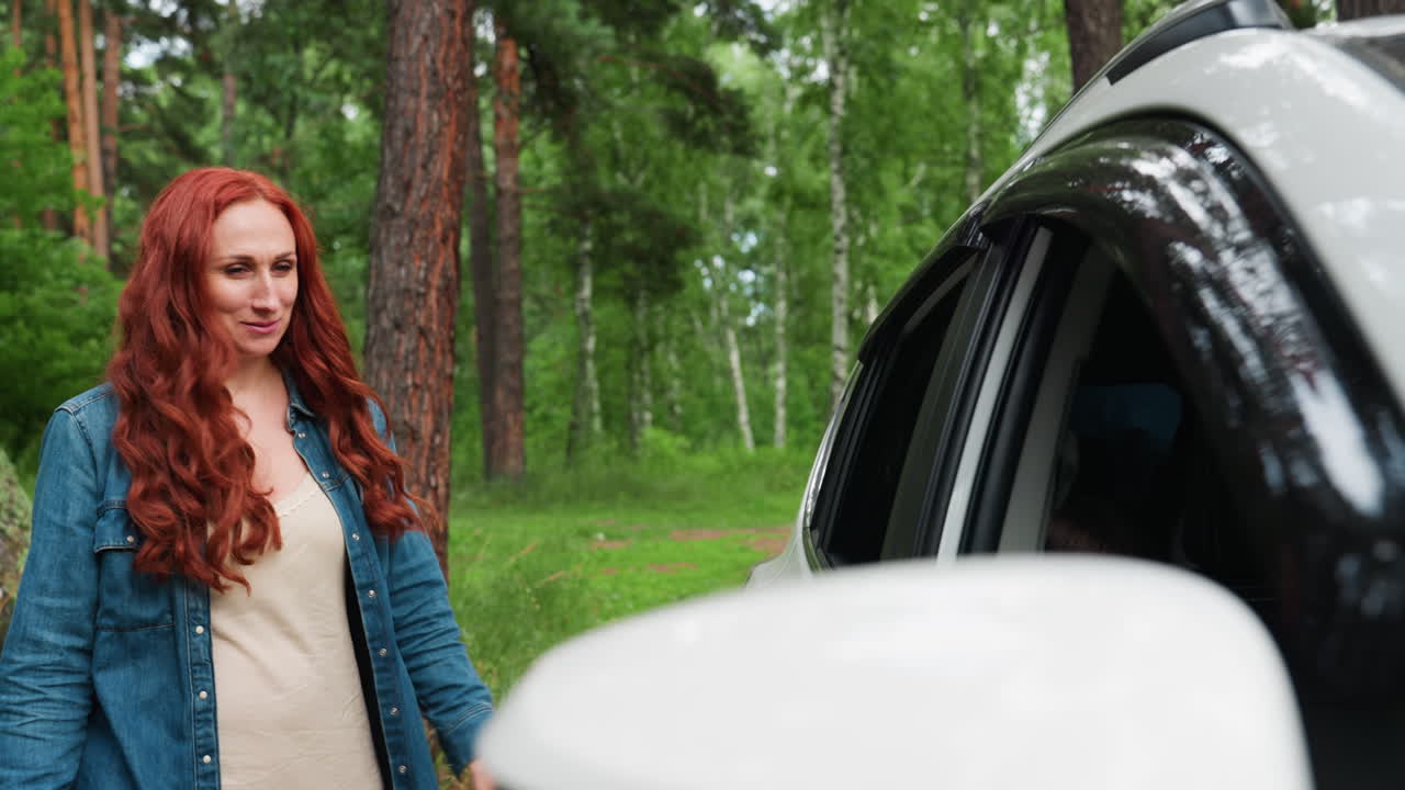 Autumn woman with long red hair smiles warmly while reaching to open door of white vehicle parked in lush arboretum, surrounded by tall trees and soft daylight, peaceful moment in nature
