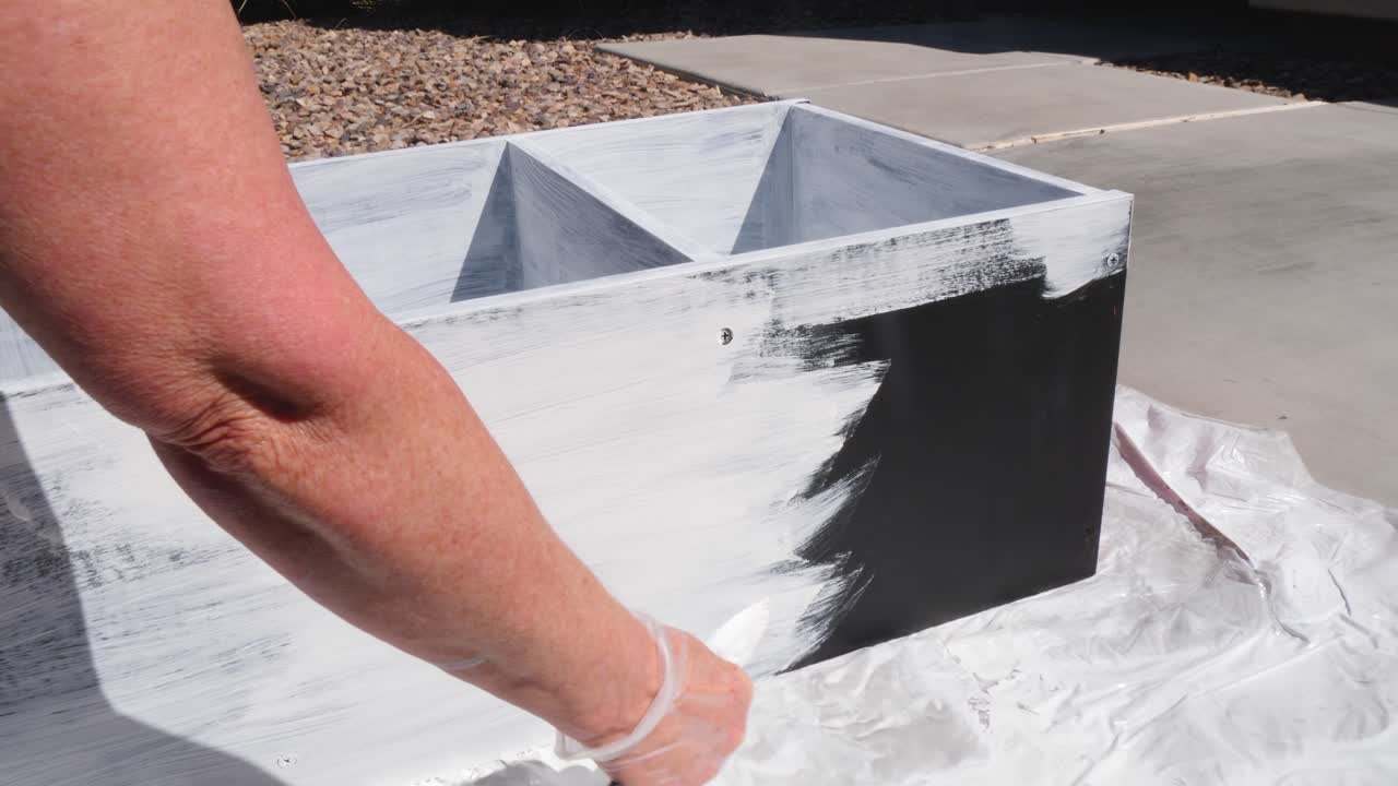 A woman paints a black cabinet white on her driveway