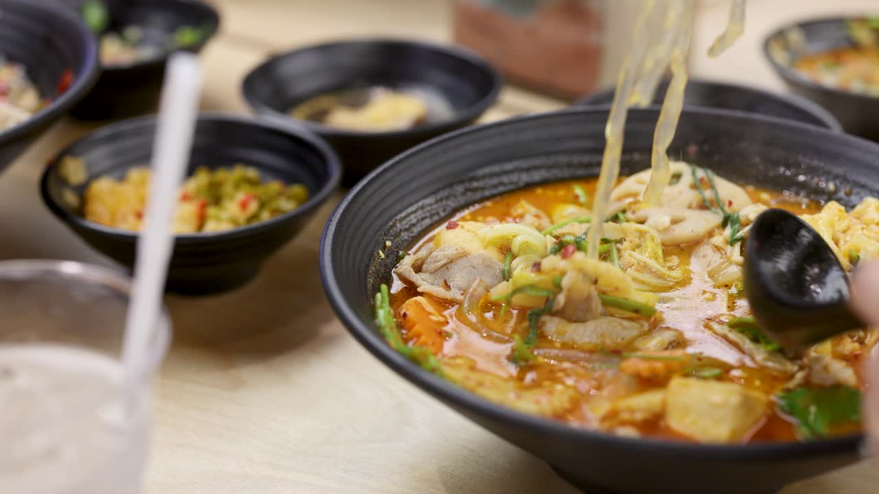 Hand lifts noodles from hearty spicy hotpot with tofu, meat, vegetables, and chili broth