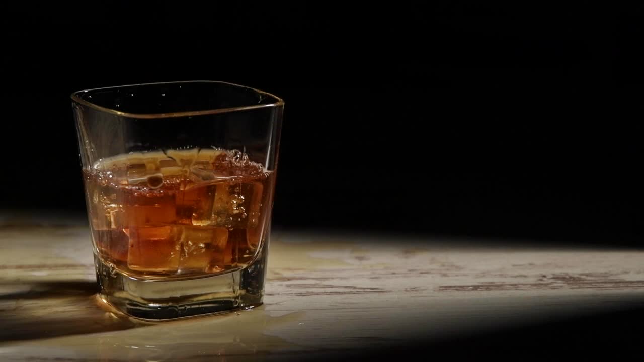 persona vertiendo cubitos de hielo en un vaso con bourbon en una mesa de madera en rayos contra un fondo negro