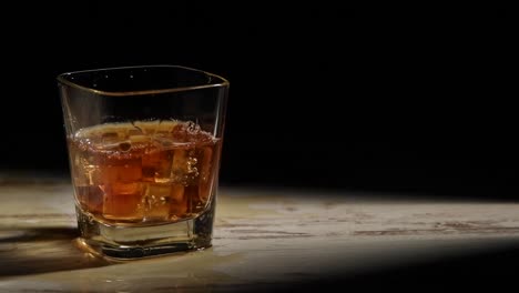 Person-pouring-ice-cubes-into-glass-with-bourbon-on-wooden-table-in-rays-against-black-background