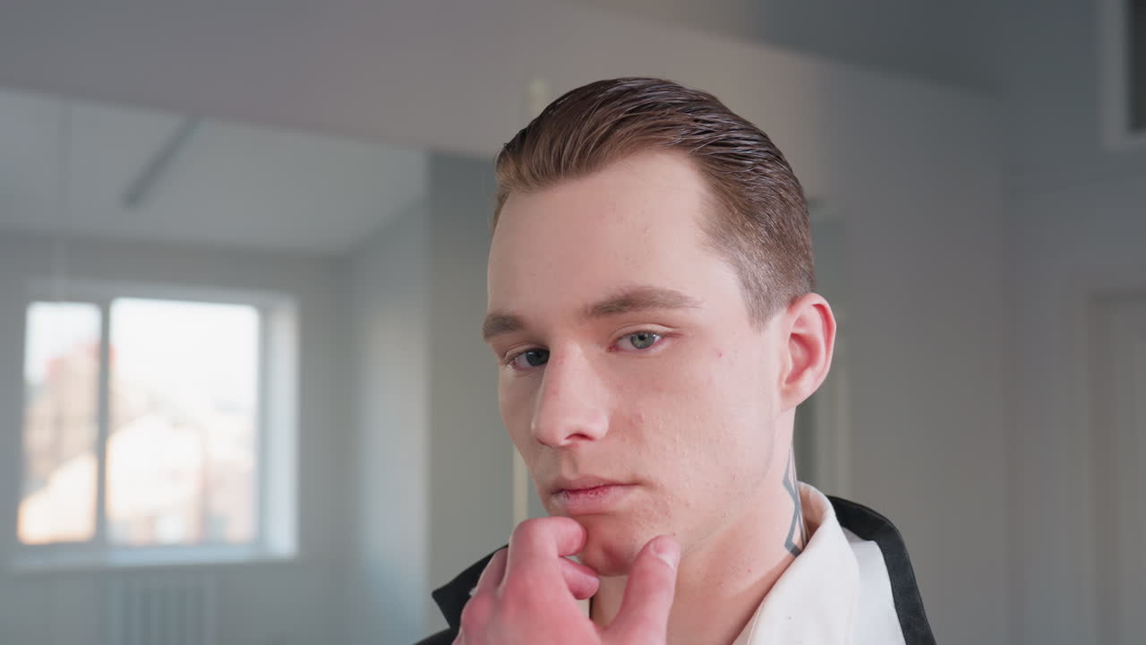 portrait view of stylish man with slicked hair and neck tattoo holding chin in thoughtful pose, standing calmly near large wall mirror reflecting soft daylight from window in minimal room interior