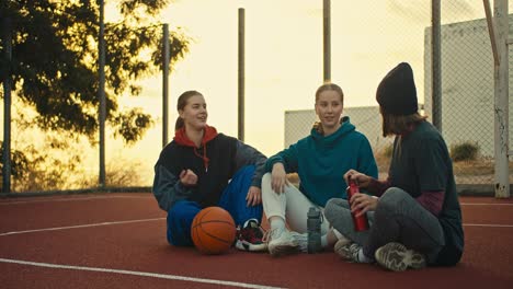 Ein-Blondes-Mädchen-In-Einer-Sportuniform-Wärmt-Sich-Auf-Und-Sitzt-Zusammen-Mit-Ihren-Freunden-Auf-Dem-Roten-Boden-Eines-Straßenbasketballplatzes-Und-Kommuniziert-Bei-Sonnenaufgang-Mit-Ihnen