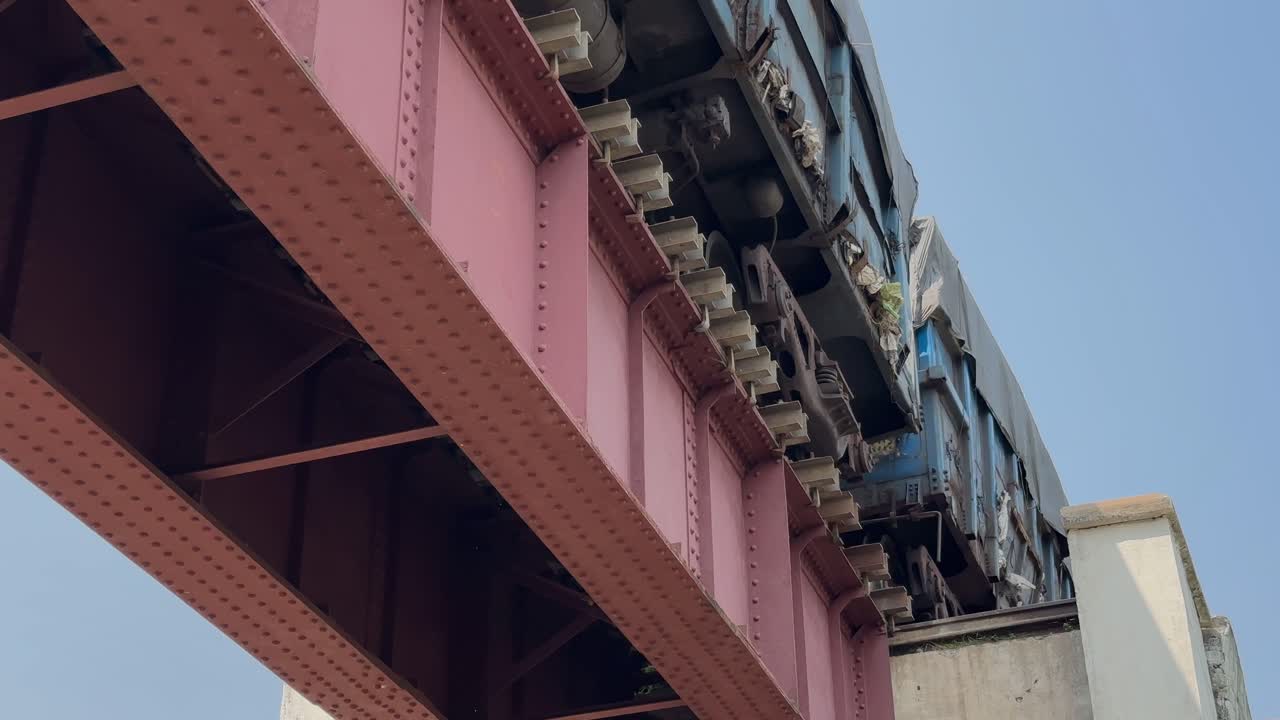 Freight wagons rumble across a steel railway bridge, captured from a low static angle showing metal beams, wheels, and vibrant industrial details