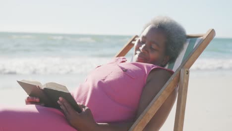 Happy-senior-african-american-woman-sitting-on-deck-chair-and-reading-book-at-beach,-in-slow-motion